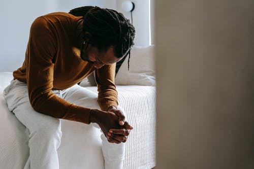 man sitting at edge of bed with elbows resting on knees and hands together with fingers laced and head down